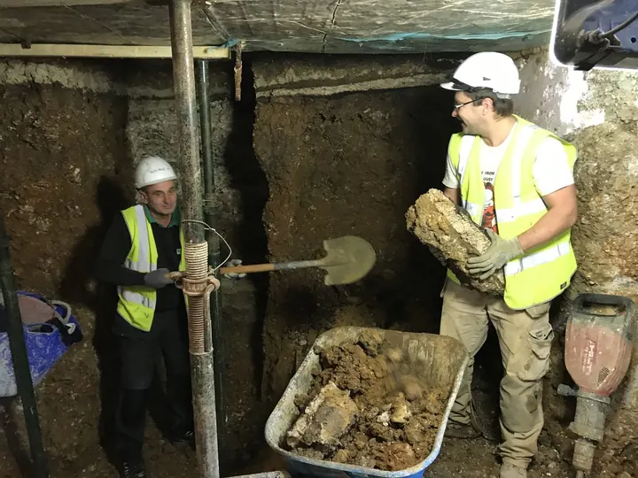 Two operatives with a spade and wheelbarrow in a hand-dug excavation under a house, supported by temporary props
