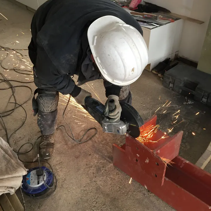 An operative in a hard hat cutting a steel beam with an angle grinder, throwing sparks